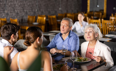 Happy senior woman and man meeting with adult children over dinner in restaurant. People talking cheerfully and drinking wine at table