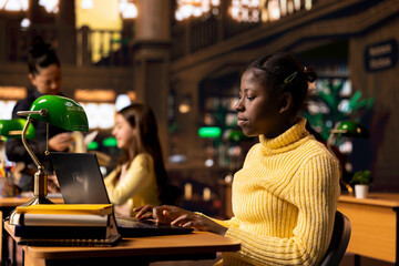 African american teen focuses on online database research at the library, works on writing an essay...