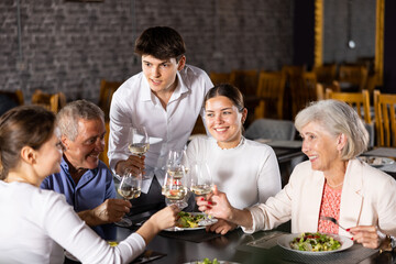 Cheerful elderly couple spending time with young friends in cozy restaurant. Man and woman having fun while talking and enjoying light dinner with wine at table