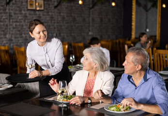 Adult woman waiter serving wine to couple of elderly man and woman in restaurant
