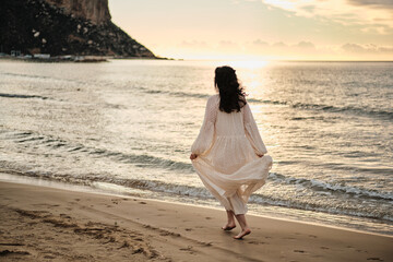 Serene woman in flowing white dress walks towards the ocean at sunset.  Peaceful beach scene evokes...