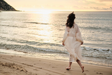 Woman in a flowing white dress walks along a sandy beach at sunset. Golden hour light illuminates...
