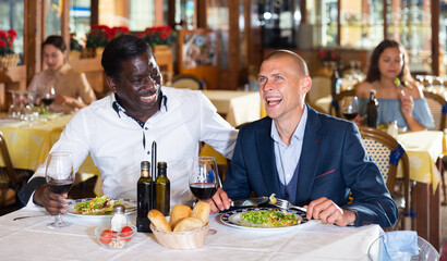 Two male colleagues enjoying dinner with wine and having conversation at restaurant