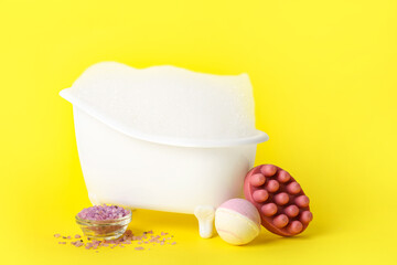 Small bathtub with foam, massage soap, bath bomb and sea salt on yellow background