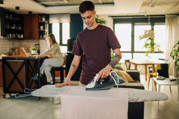 Fototapeta premium Young tattooed woman ironing clothes while her partner works on laptop in the kitchen
