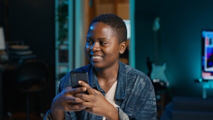 Jolly woman relaxing at home sitting at office desk, texting friends on smartphone in front of laptop. Cheerful freelancer taking break to chat with mates online using phone in living room, camera A