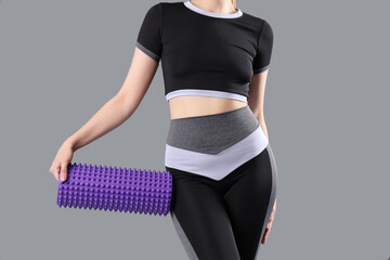 Young woman in sportswear and with foam roller on grey background