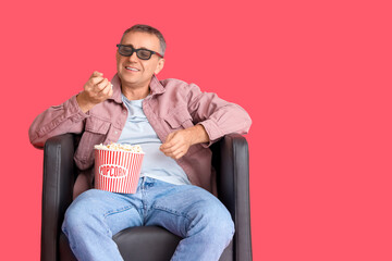 Mature man with 3D glasses and popcorn watching movie in armchair on red background