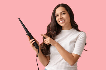 Beautiful young woman with curling iron on pink background