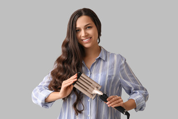 Beautiful young woman with curling iron on grey background