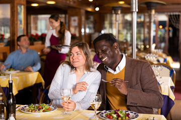 Adult african american man enjoying company of positive european woman over dinner in restaurant ..