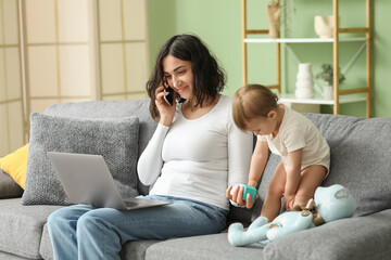 Cute baby and young mother working with laptop while talking by mobile phone on maternity leave at home