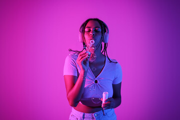 Young African-American woman with soap bubbles listening to music on neon background