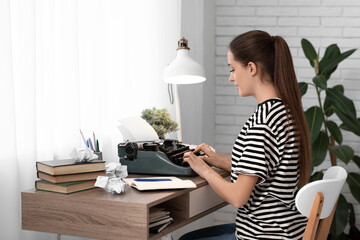 Beautiful happy female writer with typewriter books and notebook sitting at table in room