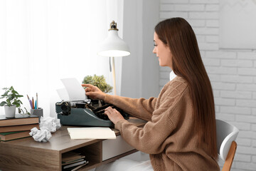 Beautiful happy female writer with typewriter and notebook sitting at table in room