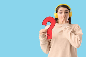 Wondering young woman in headphones with paper question mark on blue background