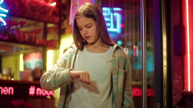 Irritated angry Asian Caucasian schoolgirl kid waiting in city annoyed teen teenager child checking time at wristwatch late evening neon impatient punctual young junior girl wait for friend meeting