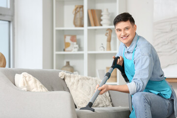 Obraz premium Young man in apron vacuum cleaning sofa at home