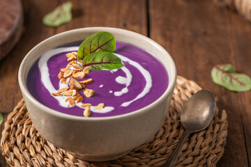Purple soup garnished with chopped nuts, a drizzle of cream, and a green leaf, served in a beige bowl on a woven mat, placed on a wooden table