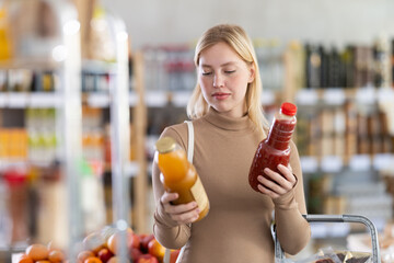 Young woman buyer chooses peach and tomato juices in bottle in grocery store