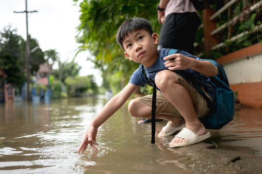Young boy crouching by floodwater, curiously touching the water while holding a backpack, symbolizing exploration.