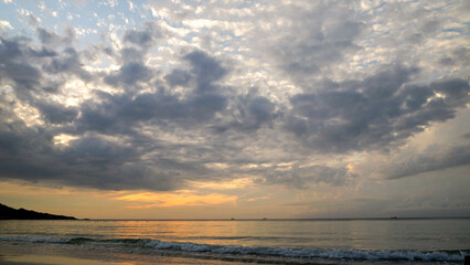 Tranquil beach scene at sunset with dramatic textured clouds covering the sky golden hues of the setting sun reflect softly on the calm ocean waves.