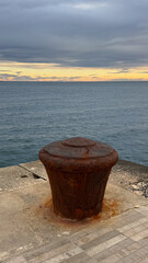 A large rusted mooring bollard on a stone pier with calm sea waters and a dramatic sunset sky in the background. 