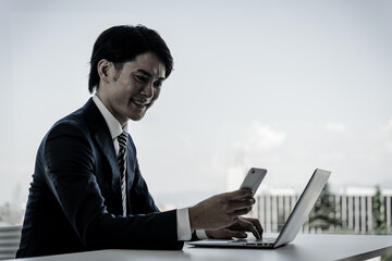 スマートフォンとノートパソコンを使いながらデスクワークする若いアジア人ビジネスマン
