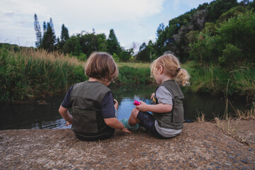 Two kids in vests nature sitting at edge of water