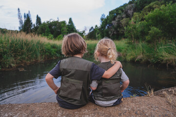 Two siblings in adventuring clothing hugging each other in front of water
