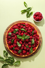 Berry pie on wooden plate, raspberries in bowl and basil leaves on light green background, top view
