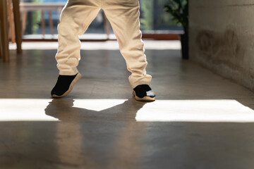 A child in beige pants and black sneakers standing indoors with sunlight casting shadows on the floor.