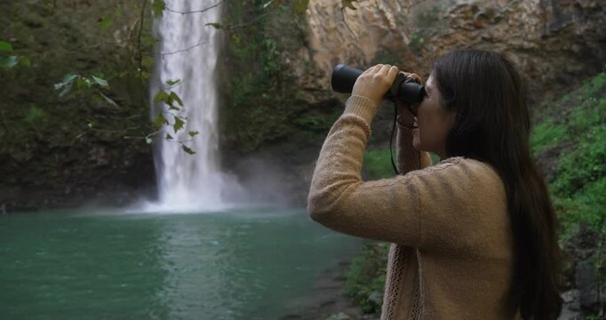 Mexican woman in the rigth side, in front of a waterfall looking at the landscape with binoculars