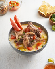 A hearty bowl of beef soup is served with chunks of tender meat, fresh tomatoes, and crispy chips, accompanied by lime and salsa on the side