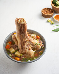A hearty bone soup features a large bone, tender chunks of meat, colorful vegetables, and fresh lime, accompanied by garnishes