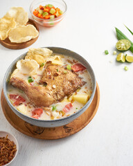 Savory soto ayam or chicken soup with vegetables sits in a bowl next to a plate of crunchy chips and a colorful fresh salad at mealtime