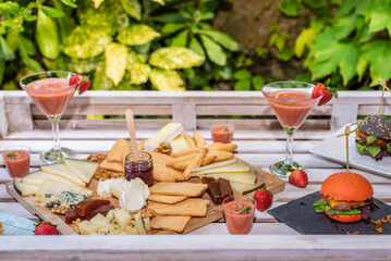A colourful assortment of cheeses, sweets and drinks on a wooden table awaits to be shared.