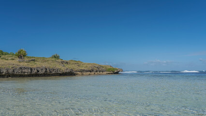 An island in the Indian Ocean. Grass and fan palm bushes grow on the rocky soil.  Crystal clear turquoise water is in the foreground.The waves are foaming in the distance. Clear blue sky. Copy space