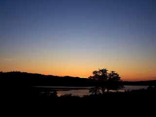 Obraz premium Sun setting over serene lake, with the warm glow of the fading light reflecting on the water and framed by a silhouette of a tree on the middle right side, serene, relaxing, scenic