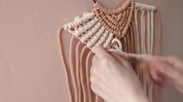 Female hands weaving macrame wall hanging with beige and brown ropes. Handmade decor piece process in slow motion. Intricate knot patterns. Bohemian charm in interior spaces.