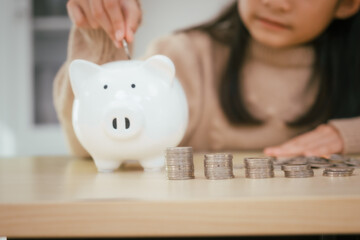 A girl sits at a table, saving money by inserting coins into a piggy bank, emphasizing the...