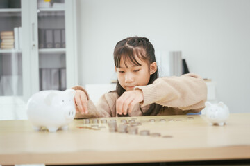 A girl sits at a table, saving money by inserting coins into a piggy bank, emphasizing the...