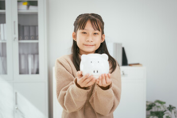 A girl sits at a table, saving money by inserting coins into a piggy bank, emphasizing the...