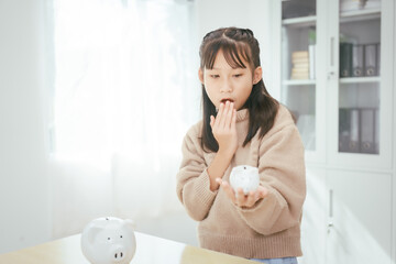 A girl sits at a table, saving money by inserting coins into a piggy bank, emphasizing the...