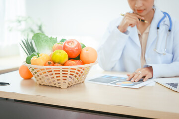 An Asian female doctor at her hospital desk works on a laptop, discussing the importance of eating fruits and vegetables for health and recommending nutritious diets online for patients' well-being
