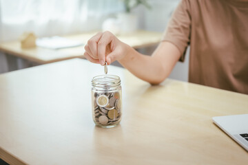 A young woman saves money for the future by putting coins in a piggy bank on the table, managing monthly expenses, and focusing on investment and business growth for financial stability