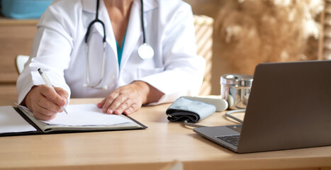 Senior woman doctor in white coat with stethoscope working with writing notes of patient while laptop on desk in modern medical office, female doctor diagnosis with concentration in clinic.