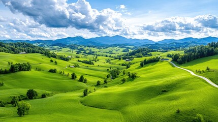 Obraz premium Lush green rolling hills and meadows stretch towards distant mountains under a cloudy blue sky.