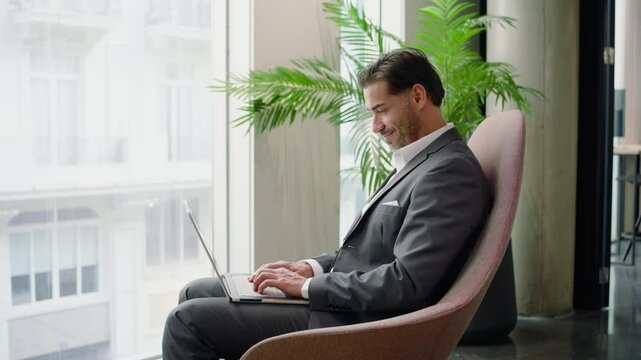 Dedicated mid age businessman in a sharp suit, diligently working on his laptop by a large window that showcases vibrant greenery outside, symbolizing the essence of contemporary remote work culture