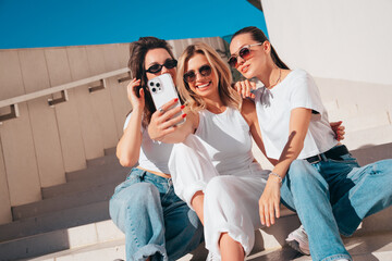 Three young beautiful smiling hipster female in trendy summer white t-shirt  and jeans clothes. Sexy carefree women posing in street. Positive models having fun, going crazy. In sunny day, take selfie